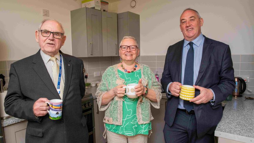 Three people standing up in a kitchen, smiling and holding mugs