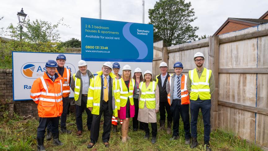 A group of people wearing hard hats and hi-vis from Sanctuary and the Renfrewshire Council stood on-site in front of signage for the 2 & 3 bedroom apartments which will be available for social rent