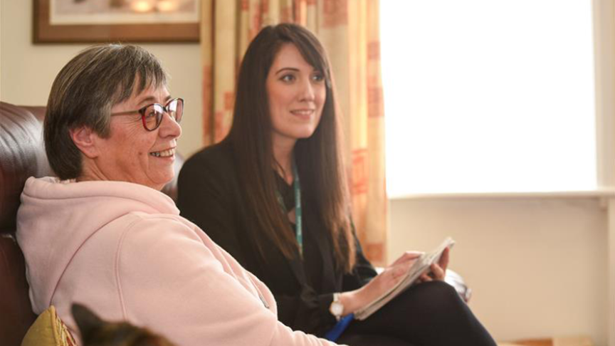 Alt text: A Sanctuary customer, a middle-aged woman with short grey hair and glasses, wearing a light pink hoodie, sits on a brown leather sofa, smiling. Next to her, a Sanctuary Housing Officer, a younger woman with long dark hair, dressed in a black blazer and wearing a lanyard, holds a notepad and pen. They are in a well-lit living room with cream curtains, a framed picture on the wall, and a cat partially visible in the foreground.