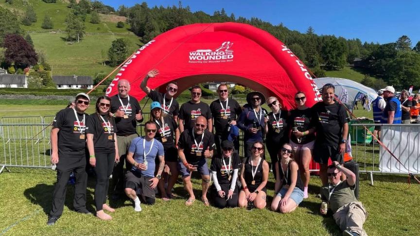 A group of people wearing black 'Walking With The Wounded' t-shirts pose in front of a red inflatable arch with the same logo. They are outdoors on grass with hills and trees in the background. Some are standing, kneeling, or sitting, and many are holding medals.
