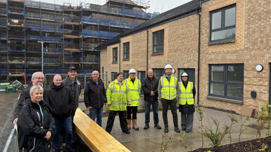 A group of men and women, some wearing hard hats and high visibility jackets, stood outside a row of new-build, brick houses. In the background is a scaffolded constructions site where more new homes are being built.