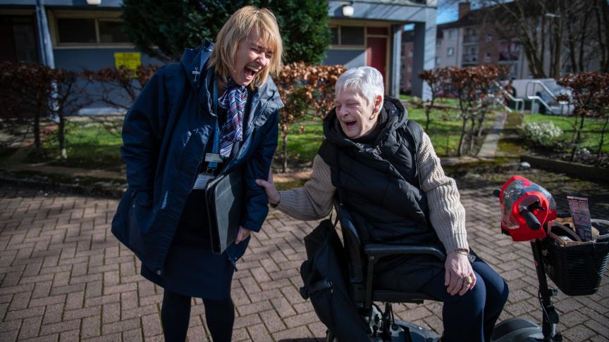 A smiling woman in a blue coat stands next to an elderly woman in a mobility scooter. They are both laughing and engaged in a joyful conversation outdoors, with a backdrop of a building and neatly trimmed bushes. 