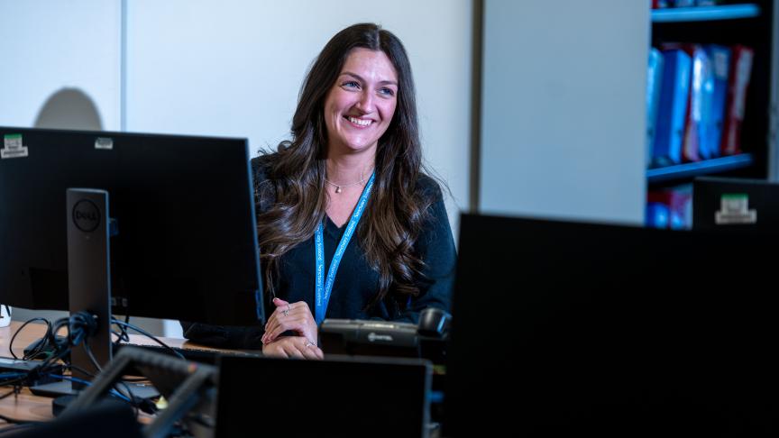 A woman with long, wavy hair is sitting at a desk in an office environment, smiling at the camera. She is positioned in front of multiple computer monitors, with a blue lanyard around her neck. 