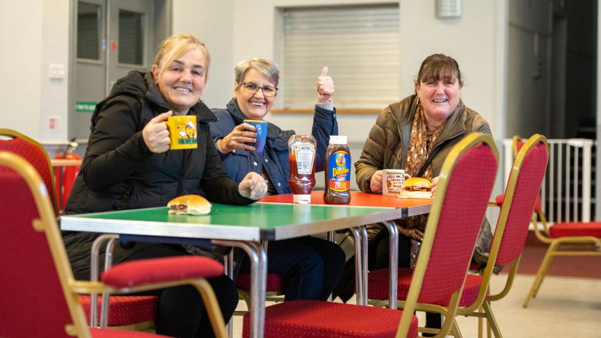 Three women are sitting at a table in a casual setting, smiling towards the camera. Each woman holds a mug, and there are bottles of sauce and sandwiches on the table.