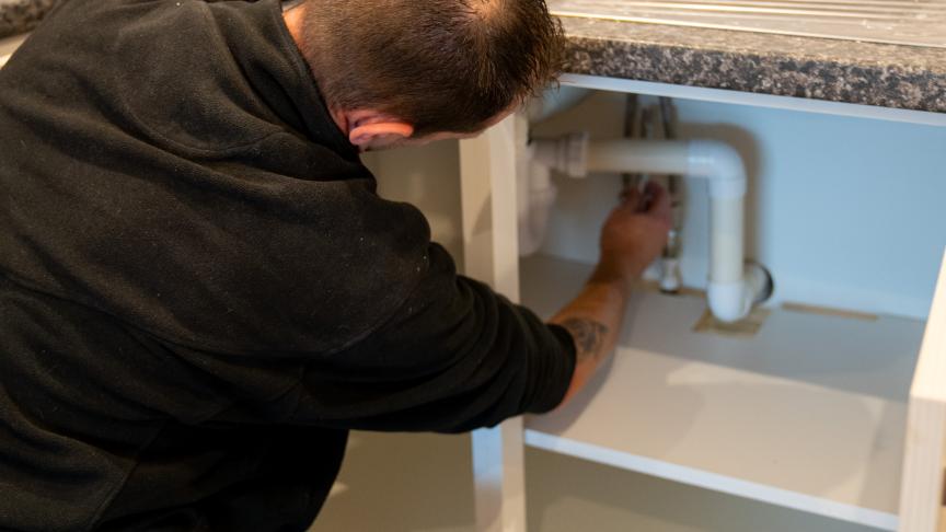 A person wearing a black hoodie and work pants is crouched down under a kitchen sink, working on the plumbing. The sink has a stainless steel surface and a faucet, and there are cleaning supplies and paint cans visible on the countertop.