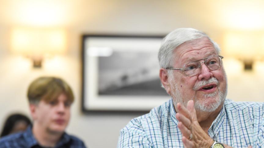 An older individual wearing glasses and a checkered shirt is seated indoors, with one hand slightly raised in a gesture as if speaking to someone out of frame. The person also wears a wristwatch on the left wrist. In the background, another individual is visible but blurred, appearing to listen to the conversation.