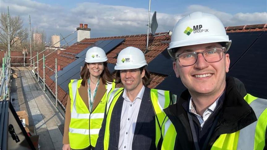 Donna Williams, Group Director of Strategy and Sustainability, is pictured with colleagues during a rooftop site visit. The group is standing on a scaffolded roof fitted with rows of solar panels, wearing hard hats and high visibility vests. Residential rooftops and a partly cloudy sky are visible in the background.