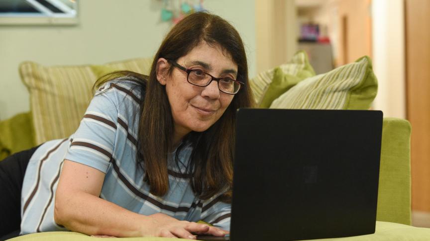 A woman with long brown hair and glasses is lying on a green couch, intently looking at a laptop in front of her. The background features a softly lit room with a hint of decor, suggesting a cozy home environment.