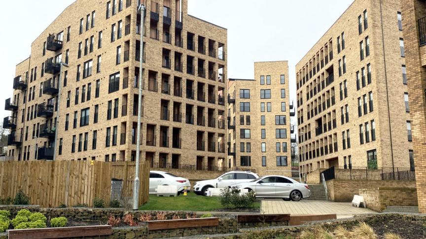Modern mid-rise apartment buildings built with light brick surround a landscaped courtyard with terraced stone planters, benches, and small shrubs. Several parked cars sit on a grassy area and paved drive between the buildings under an overcast sky