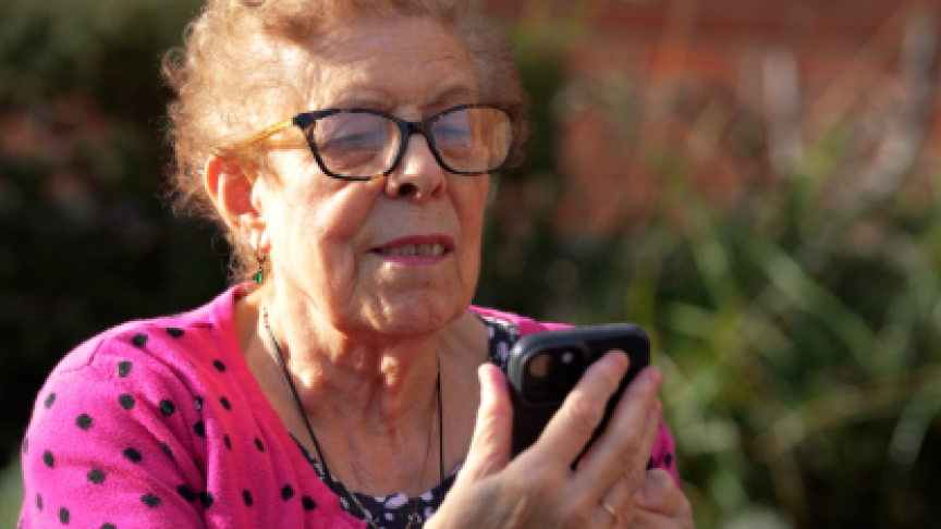 An older woman who is wearing glasses is sat outside holding a smartphone and looking at the screen, appearing to be reading something. 