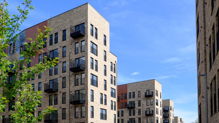 Modern mid-rise apartment buildings with light brick facades and black metal balconies line a residential development under a bright blue sky. Green leafy shrubs fill the foreground, while multiple contemporary buildings create a courtyard-like streetscape in the background.