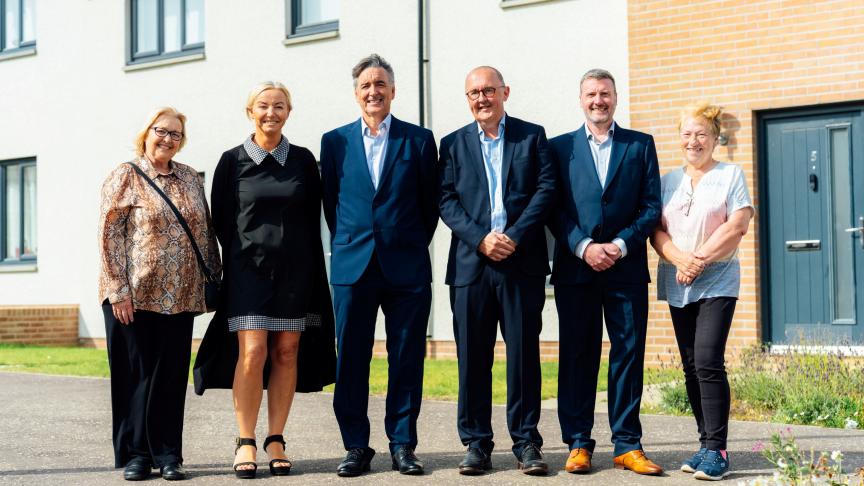 Six adults stand side by side on a paved pathway outside a modern residential development. Three people wear dark business suits, while the others wear casual clothing. The group is positioned in front of a newly built apartment or housing block with white walls, brick detailing, and large windows. A dark blue front door marked with the number 5 is visible on the right, with grass and planting beds surrounding the building in bright daylight.