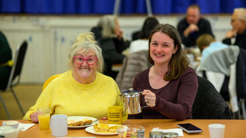 A cheerful scene in a community hall shows two women sitting at a table. The older woman on the left is wearing a bright yellow sweater and pink glasses, smiling warmly. The younger woman on the right, dressed in a dark top, is also smiling and holding a metal teapot. The table is set with plates of food, a yellow mug, and a glass of juice. In the background, other people are seated at tables, engaged in conversation.