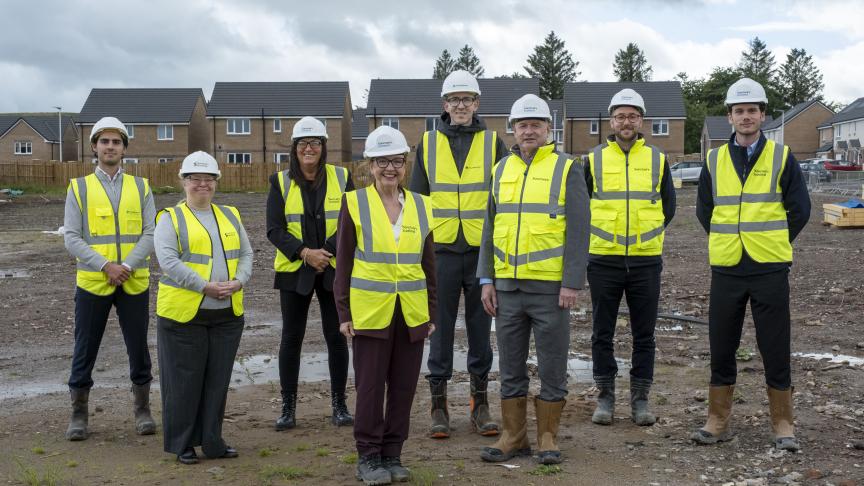 Eight people wearing white hard hats, high-visibility yellow safety vests and work boots stand on a muddy housing construction site. Newly built brick houses line the background under a cloudy sky. The group faces the camera in front of the developing residential area, with construction materials and vehicles visible around the site.