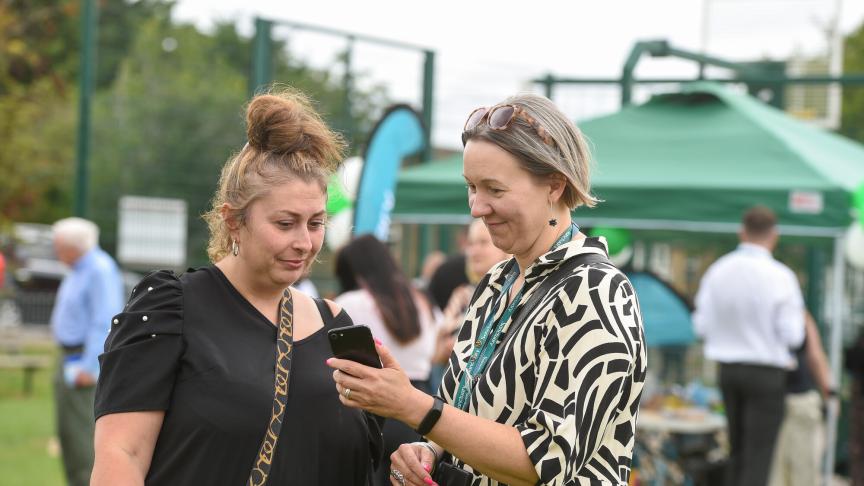 Two women are standing outdoors, engaged in conversation while looking at a smartphone. One woman, with a bun hairstyle and wearing a black top, appears to be listening intently. The other woman, with short grey hair and dressed in a patterned black and white outfit, is showing something on the phone. In the background, there are people mingling and green tents set up, indicating a social event or gathering.
