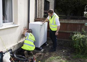 Two white, male Sanctuary staff members wearing high vis jackets are outside a building fitting a swaffham heat pump