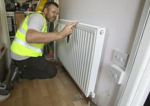 A man wearing a yellow high visibility jacket and protective goggles kneeling in a hallway working on a radiator