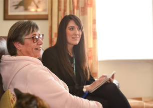 Alt text: A Sanctuary customer, a middle-aged woman with short grey hair and glasses, wearing a light pink hoodie, sits on a brown leather sofa, smiling. Next to her, a Sanctuary Housing Officer, a younger woman with long dark hair, dressed in a black blazer and wearing a lanyard, holds a notepad and pen. They are in a well-lit living room with cream curtains, a framed picture on the wall, and a cat partially visible in the foreground.