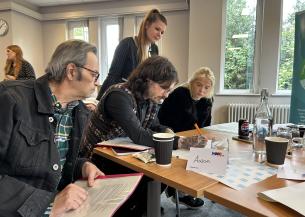 Sanctuary customers seated around a table during a training session on supporting the organisation with complaints. One person stands assisting others. The table has papers, drinks, and a name card reading 'Aaron'. The room is bright with large windows