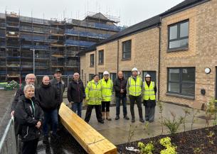 A group of men and women, some wearing hard hats and high visibility jackets, stood outside a row of new-build, brick houses. In the background is a scaffolded constructions site where more new homes are being built.