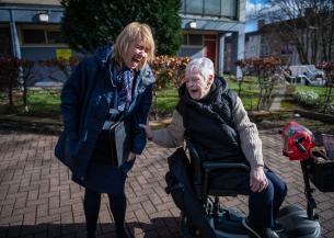 A smiling woman in a blue coat stands next to an elderly woman in a mobility scooter. They are both laughing and engaged in a joyful conversation outdoors, with a backdrop of a building and neatly trimmed bushes. 