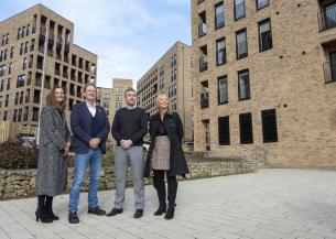 Four people standing in an outdoor courtyard area surrounded by modern multi-storey brick apartment buildings, with landscaped stone planters and paved walkways
