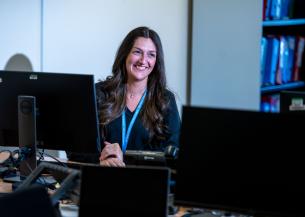 A woman with long, wavy hair is sitting at a desk in an office environment, smiling at the camera. She is positioned in front of multiple computer monitors, with a blue lanyard around her neck. 