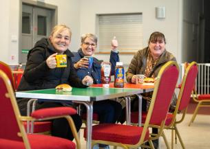 Three women are sitting at a table in a casual setting, smiling towards the camera. Each woman holds a mug, and there are bottles of sauce and sandwiches on the table.