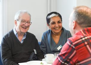 Three people sit around a small table, engaged in conversation with warm smiles. Cups and plates are on the table in a bright indoor setting.