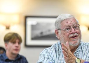 An older individual wearing glasses and a checkered shirt is seated indoors, with one hand slightly raised in a gesture as if speaking to someone out of frame. The person also wears a wristwatch on the left wrist. In the background, another individual is visible but blurred, appearing to listen to the conversation.