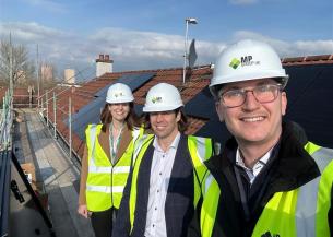 Donna Williams, Group Director of Strategy and Sustainability, is pictured with colleagues during a rooftop site visit. The group is standing on a scaffolded roof fitted with rows of solar panels, wearing hard hats and high visibility vests. Residential rooftops and a partly cloudy sky are visible in the background.