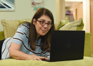 A woman with long brown hair and glasses is lying on a green couch, intently looking at a laptop in front of her. The background features a softly lit room with a hint of decor, suggesting a cozy home environment.