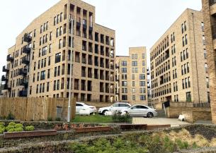 Modern mid-rise apartment buildings built with light brick surround a landscaped courtyard with terraced stone planters, benches, and small shrubs. Several parked cars sit on a grassy area and paved drive between the buildings under an overcast sky