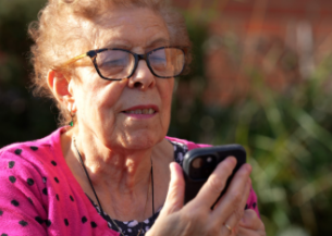 An older woman who is wearing glasses is sat outside holding a smartphone and looking at the screen, appearing to be reading something. 