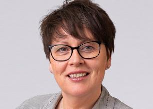 Head-and-shoulders portrait of Kathryn Fox-Rogers wearing glasses and a light grey jacket against a grey background.
