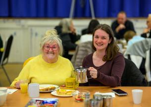 A cheerful scene in a community hall shows two women sitting at a table. The older woman on the left is wearing a bright yellow sweater and pink glasses, smiling warmly. The younger woman on the right, dressed in a dark top, is also smiling and holding a metal teapot. The table is set with plates of food, a yellow mug, and a glass of juice. In the background, other people are seated at tables, engaged in conversation.