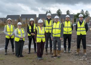 Eight people wearing white hard hats, high-visibility yellow safety vests and work boots stand on a muddy housing construction site. Newly built brick houses line the background under a cloudy sky. The group faces the camera in front of the developing residential area, with construction materials and vehicles visible around the site.