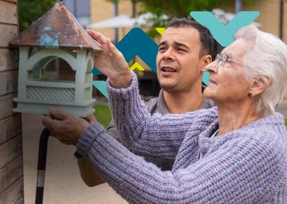 An elderly woman is helped by a younger man to hang a green bird house on a wooden wall