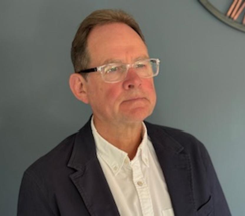 Person in a dark blazer over a white button-up shirt standing against a plain background, with part of a clock visible in the top right corner.
